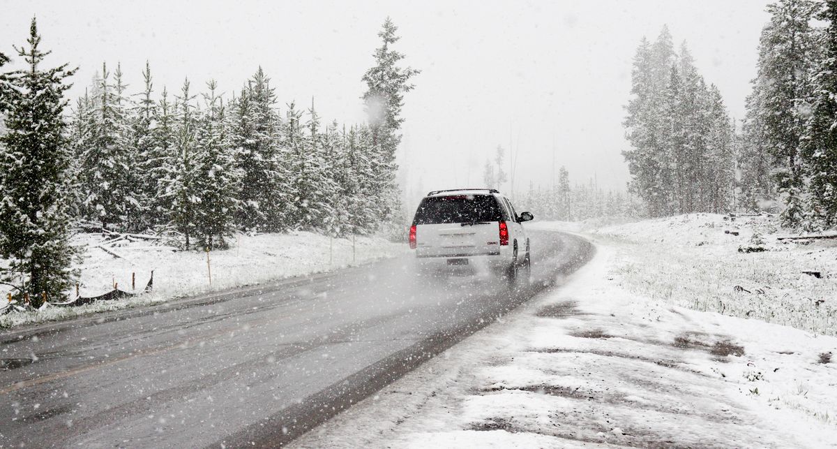 A car drives on a snowy road surrounded by trees during a snowstorm