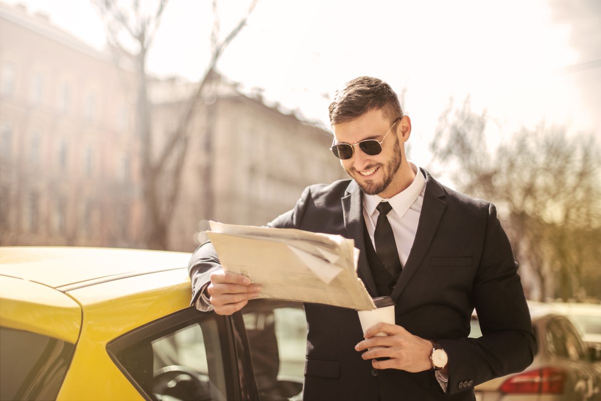 Smiling businessman in a suit reads a newspaper next to a yellow car