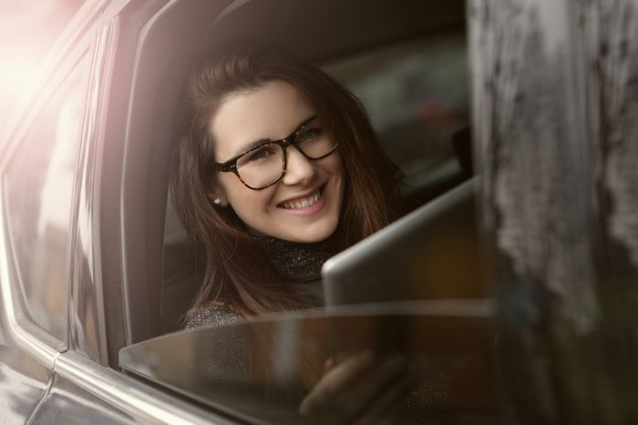 A cheerful young woman with glasses smiles while looking out of a car window