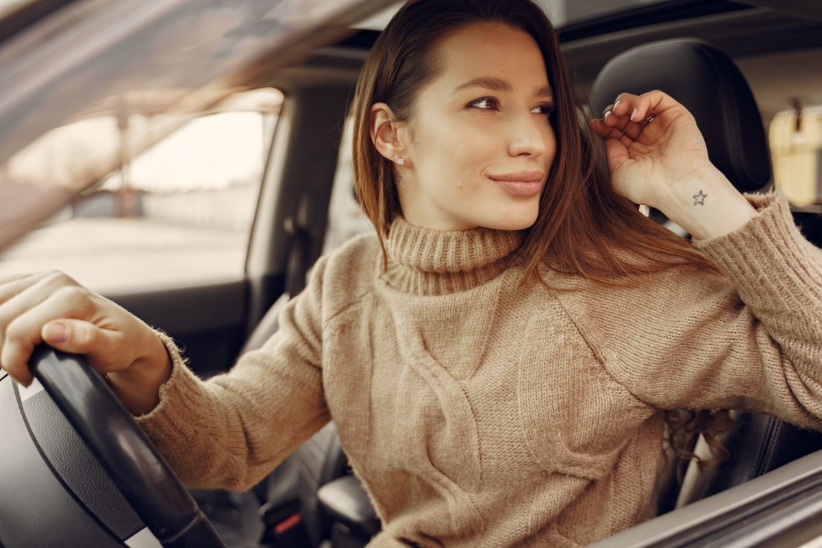 Positive young female driver in warm clothes at wheel of leather interior car going around city streets while greeting friend during riding