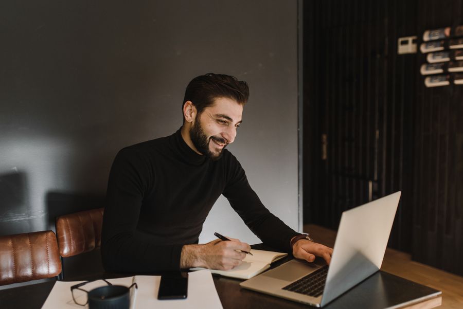 Man with beard smiling while working on a laptop in contemporary office setting