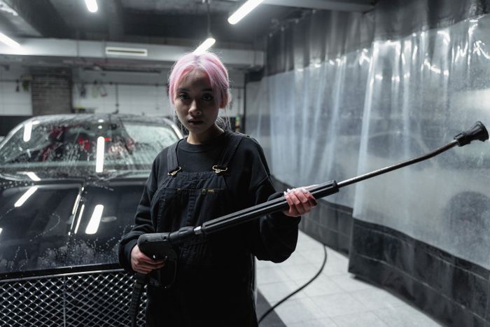 A woman with pink hair uses a pressure washer in a car wash setting