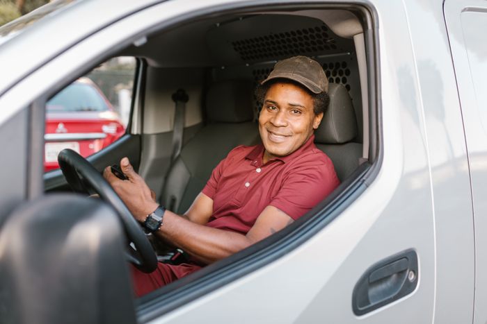 A smiling delivery driver in uniform seated inside his van, holding a smartphone