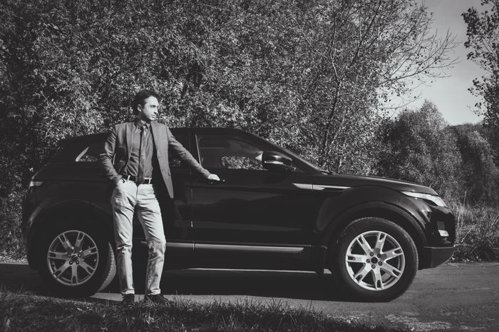 Man in formal attire stands confidently by a black SUV in a natural setting