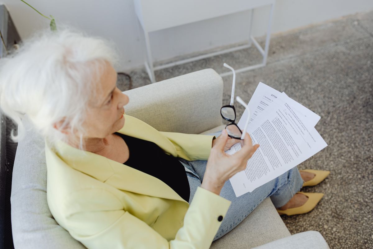 Elderly woman in a blazer holding glasses and documents, sitting indoors
